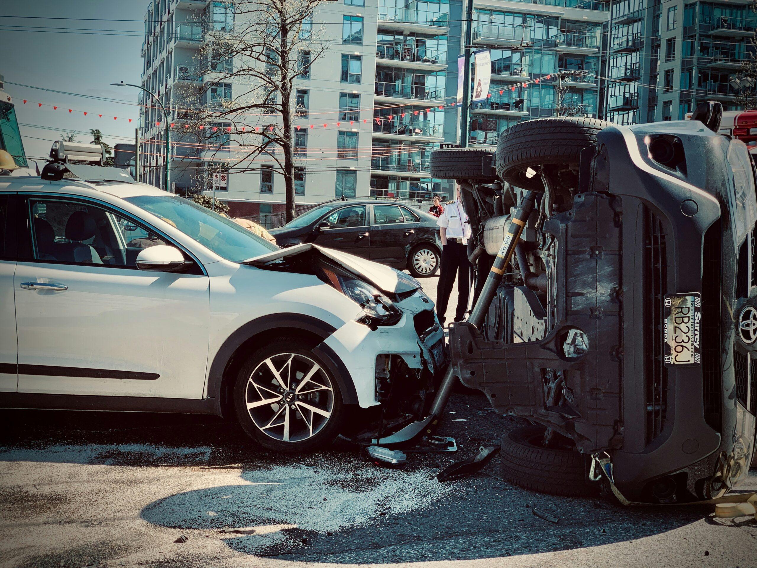 Scene of a serious car accident involving two vehicles — one flipped on its side and another with a crushed front, in an urban street setting with police officers inspecting the crash site