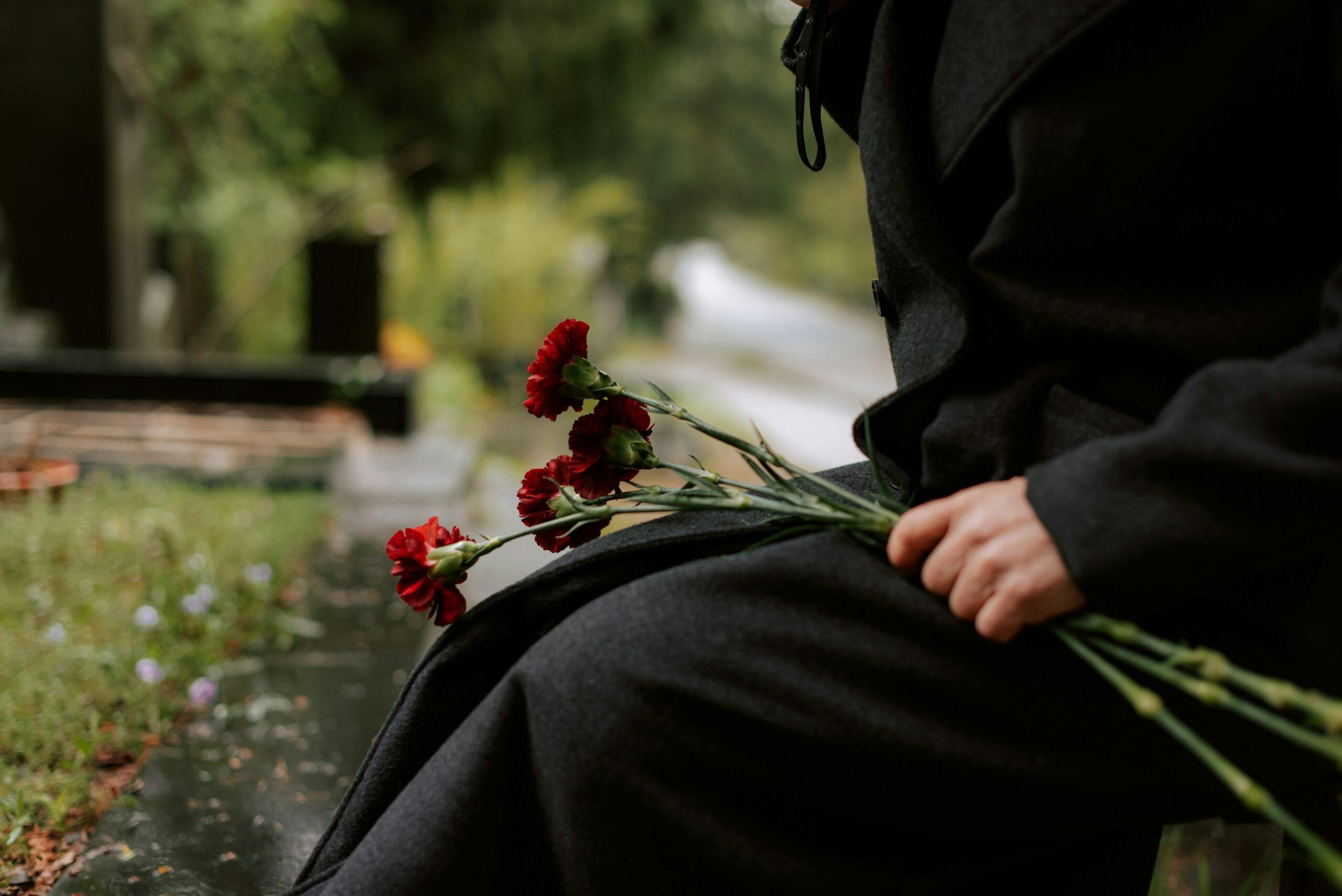 Person dressed in black holding red carnations while sitting at a grave, symbolizing grief and remembrance after loss