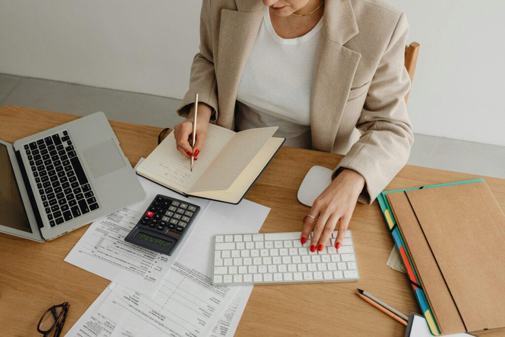 A person working at a desk with a laptop, calculator, tax documents, and folders, writing notes in an open notebook with one hand while typing on a wireless keyboard with the other, in a clean office setting.