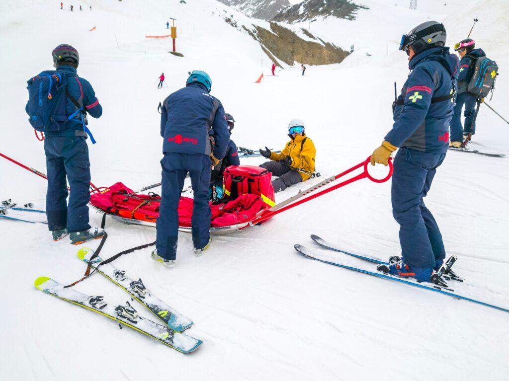 A group of ski patrollers tending to an injured skier on a snowy mountain slope, with rescue equipment, a red rescue sled, scattered skis, and other skiers in the background.