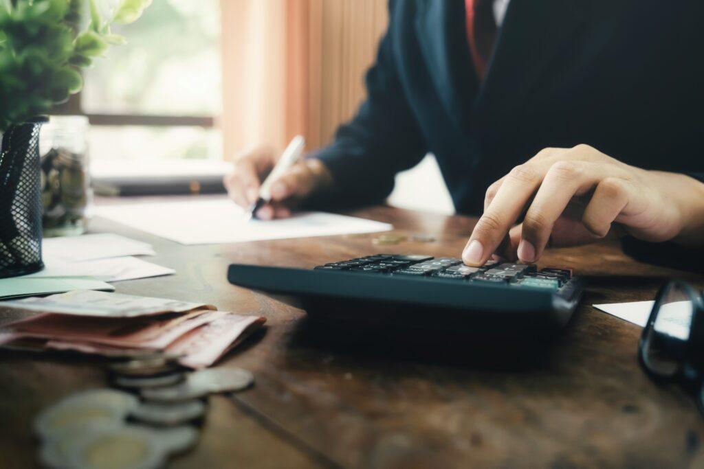 Man in a dark suit working at a wooden desk, pressing buttons on a calculator with one hand while writing on paperwork with the other, with scattered coins, cash, and office supplies in the foreground.