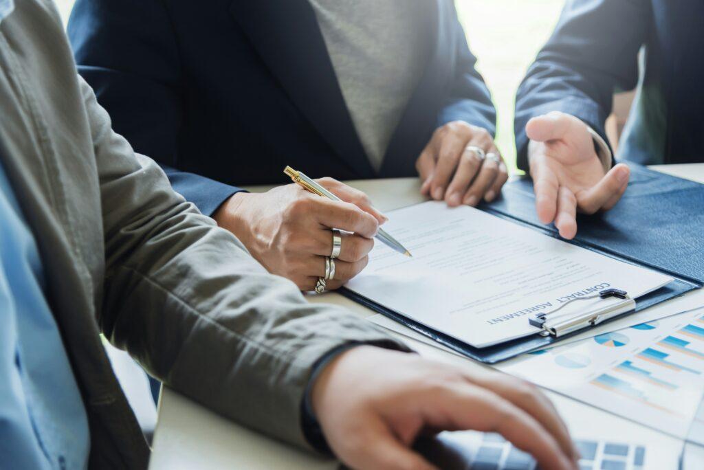 A close-up view of three people seated at a table reviewing and signing a contract, with one person holding a pen over the document, another gesturing with their hand, and financial charts visible in the foreground.