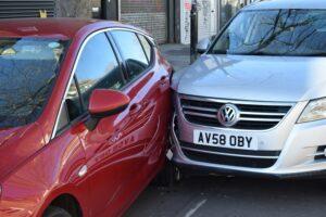 A close-up view of a collision between two parked cars on a city street, with a red hatchback showing a deep dent along its side where a silver Volkswagen has impacted it, both vehicles pressed tightly together near the front doors, with reflections of nearby buildings visible on the car surfaces.