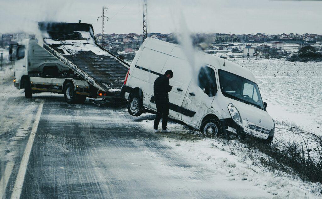 A white cargo van partially off the road and tilted into a snowy ditch while being recovered by a flatbed tow truck on an icy rural roadway, with a man standing beside the van during winter conditions and light snowfall.