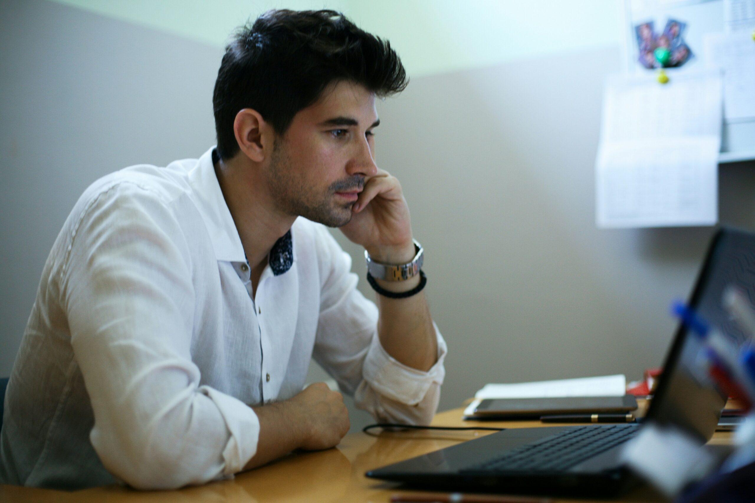 A man seated at a desk, focused on his laptop, with a notepad and pen beside him.