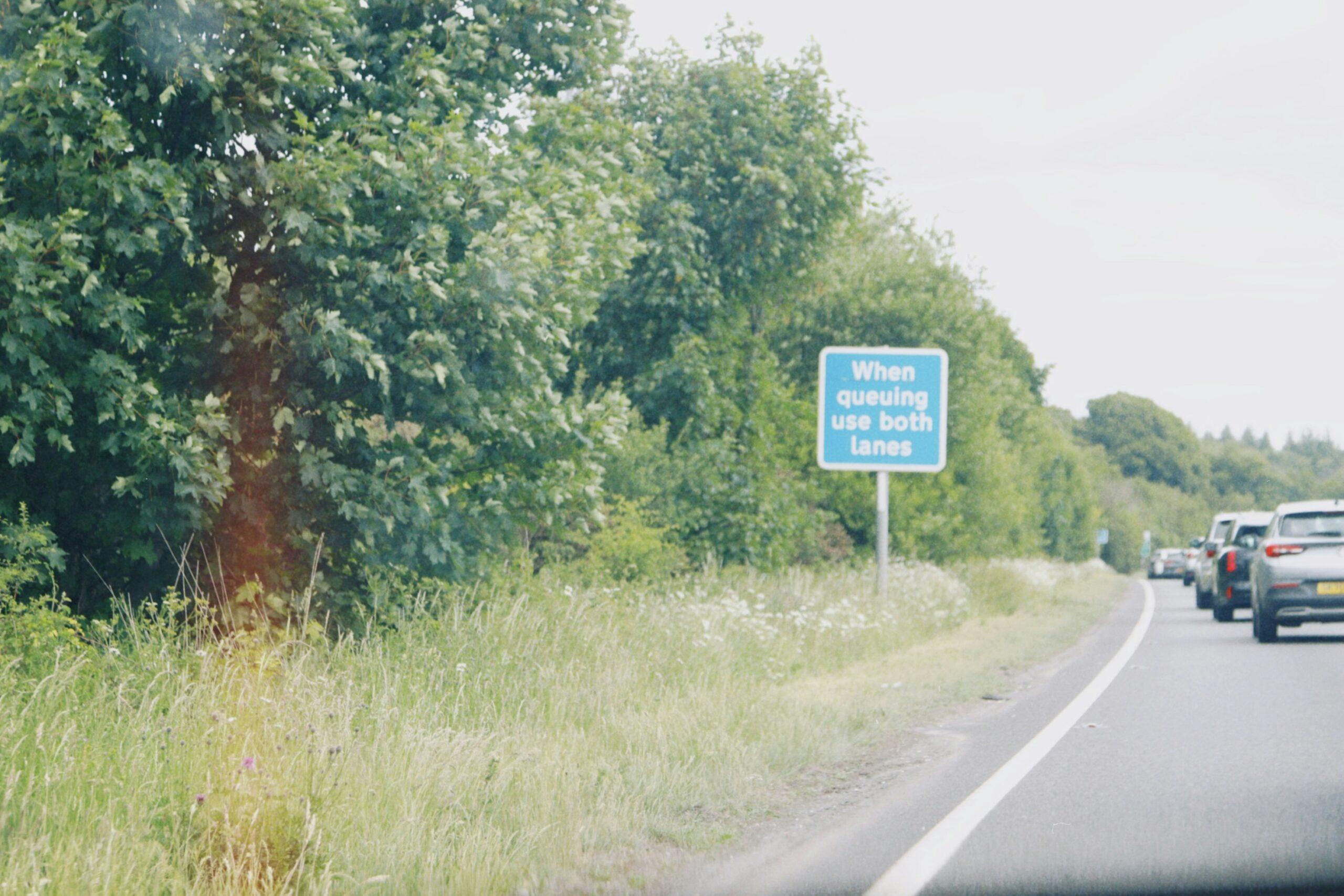 A line of cars queued along a two-lane roadway bordered by tall green trees and wild grass, with a blue roadside sign reading “When queuing use both lanes,” under an overcast sky, viewed from the shoulder of the road as traffic slows ahead.
