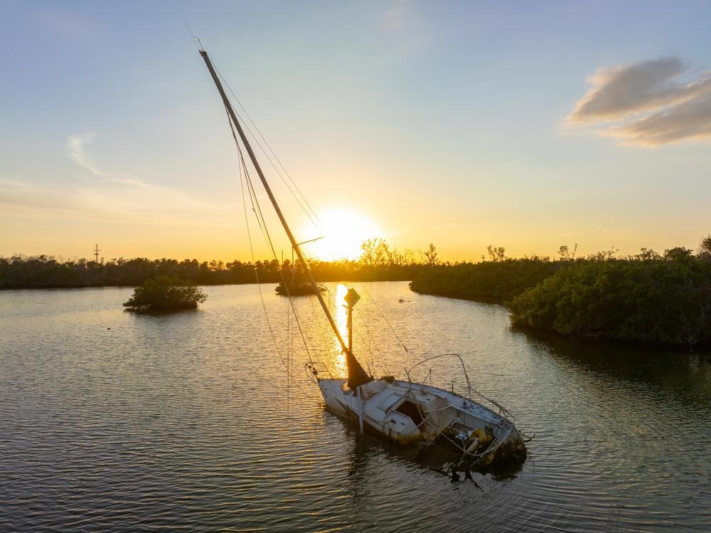 A partially sunken sailboat leans in calm water at sunset, surrounded by mangroves.