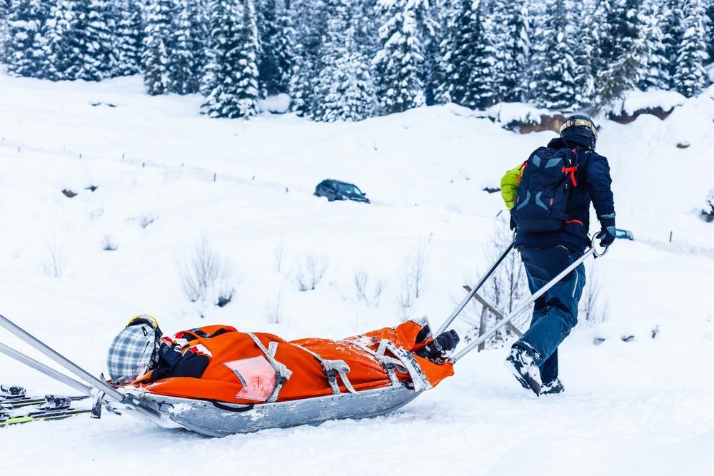 A ski patroller pulls an injured skier strapped into a rescue sled through the snow toward a road.