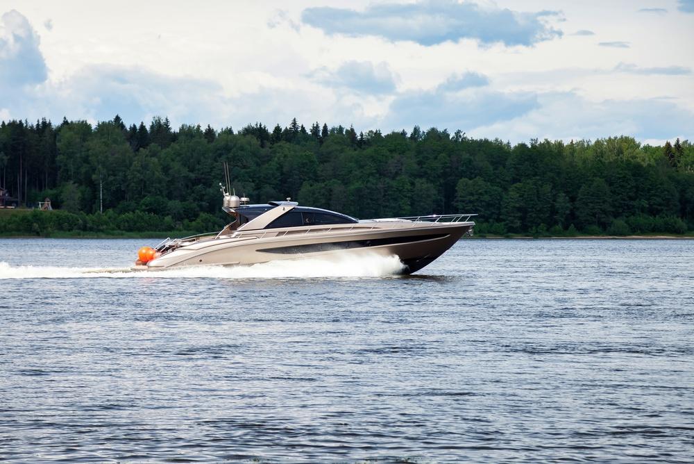 A sleek speedboat cruises across a lake in front of a dense tree line under a partly cloudy sky.
